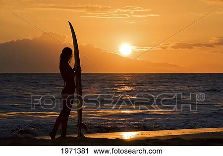 Stock Image - Hawaii, Female Surfer On Beach Silhouetted Against Orange Sunset Over Ocean.. Fotosearch