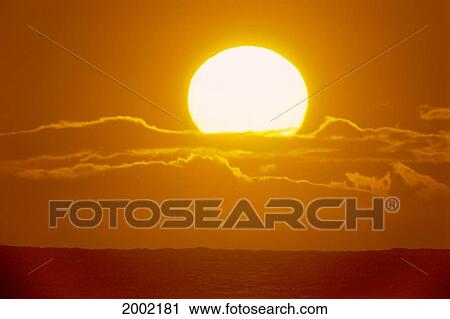 Stock Image - Hawaii, Glowing Sun Ball Partially Covered With Clouds In Bright Orange Skies Over Ocean. Fotosearch