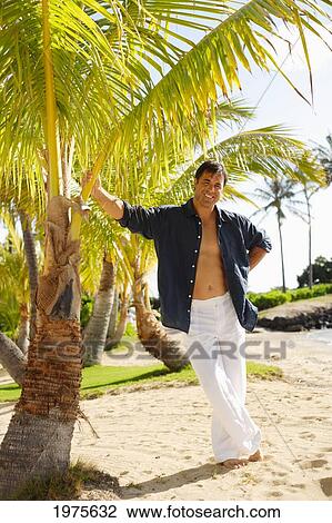 Stock Image - Hawaii, Handsome Man Standing On Beach Next To Palm Tree.. Fotosearch
