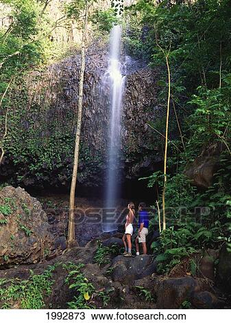 Stock Image - Hawaii, Kauai, Couple Looking Up At Waterfall.. Fotosearch