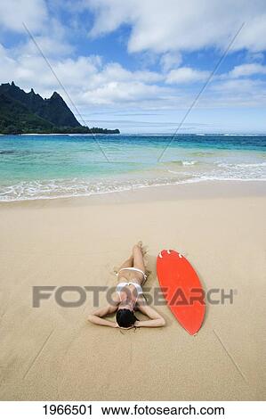 Stock Image - Hawaii, Kauai, Haena Beach Tunnels Beach, Woman Laying On Beach With Surfboard.. Fotosearch