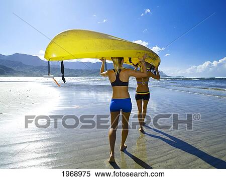 Stock Photography - Hawaii, Kauai, Hanalei Bay, Two Active Young Woman Carrying A Kayak Into The Ocean.. Fotosearch