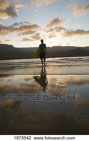 Stock Image - Hawaii, Kauai, Hanalei Bay, Young Man At The Beach Walking With Surfboard At Sunset.. Fotosearch