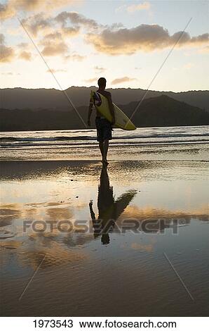 Stock Image - Hawaii, Kauai, Hanalei Bay, Young Man At The Beach Walking With Surfboard At Sunset.. Fotosearch