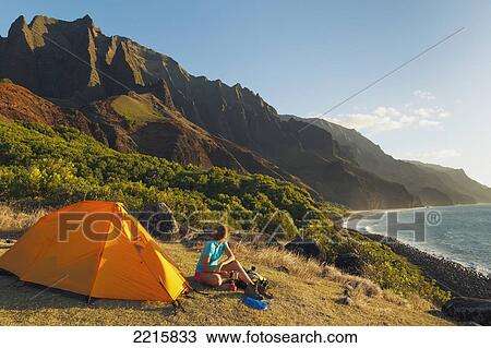 Stock Image - Hawaii, Kauai, Na Pali Coast, View of the Kalalau Mountains and ocean, Female camper enjoys the scenery.. Fotosearch