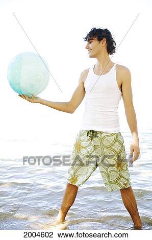 Stock Image - Hawaii, Kauai, North Shore, Handsome Guy Holds Ball While Standing In Ocean.. Fotosearch