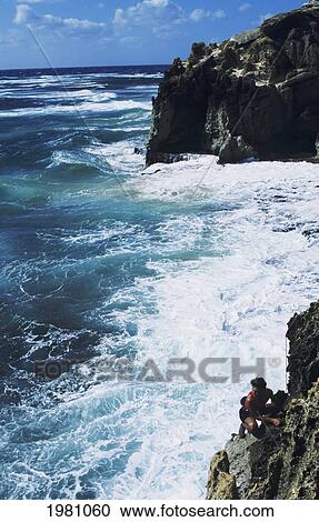 Stock Image - Hawaii, Kauai, Poipu, Man Rock Climbing Along Rocky Coastline, Turbulent Ocean Below. Fotosearch