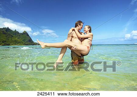 Stock Image - Hawaii, Kauai, Tunnels Beach, a young man holding a beautiful girl in the water.. Fotosearch