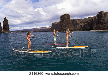 Stock Image - Hawaii, Lanai, Three young friends stand-up paddle boarding off the coast.. Fotosearch