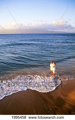 Stock Photo - Hawaii, Maui, Full Length View Of Senior Couple Walking Along Shore Line At Sunset, Golden Light. Fotosearch