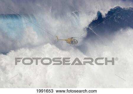 Stock Image - Hawaii, Maui, Helicopter Filming Tow-In Surfer At Peahi Aka Jaws, Wild Crashing Waves.. Fotosearch