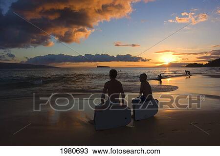Stock Image - Hawaii, Maui, Makena State Park, Oneloa Or Big Beach, Two Kids Sit On Boogieboards Watching A Dramatic Sunset.. Fotosearch