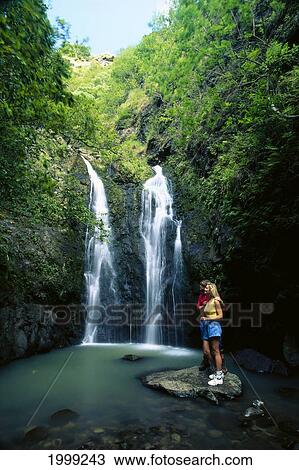 Stock Image - Hawaii, Maui, Mokumokuele Valley And Rainforest, Young Couple At Foot Of Waterfall. Fotosearch