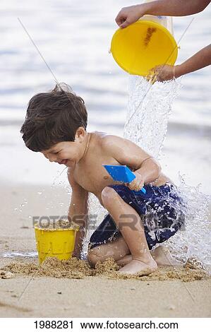 Stock Image - Hawaii, Maui, Spreckelsville, Baby Beach, Young Boy Playing In Sand Gets A Bucket Of Water Poured On Him.. Fotosearch