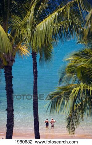 Stock Image - Hawaii, Maui, Wailea, Mokapu Beach, Senior Couple Stands Knee Deep In Water Looking Out At Ocean, Palm Trees Surround.. Fotosearch