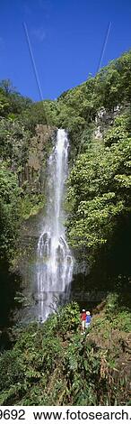 Stock Image - Hawaii, Maui, Wailua Falls, Couple Look At Waterfall, Foliage, Blue Sky Panoramic C1640. Fotosearch