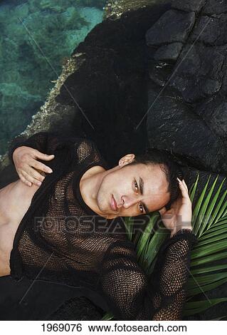 Stock Photograph - Hawaii, Oahu, Attractive Male Wearing A Net Shirt While Laying On Rocks And Palm. Fotosearch