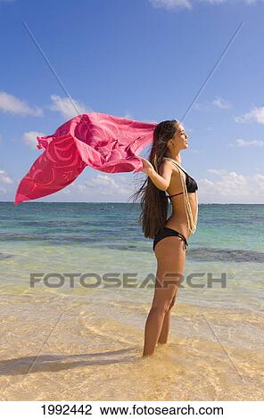 Stock Image - Hawaii, Oahu, Beautiful Local Girl Standing In The Ocean At A Tropical Beach, Holding A Pink Pareo.. Fotosearch