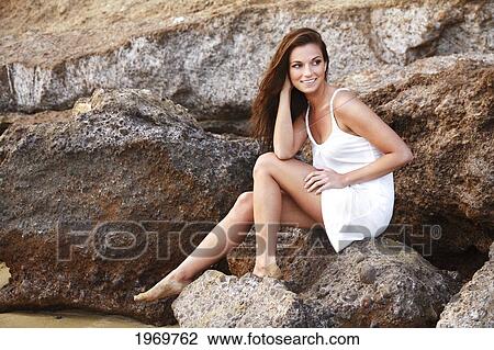 Stock Image - Hawaii, Oahu, Beautiful Woman Smiling In A White Dress Sitting On Rocks.. Fotosearch