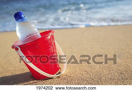 Stock Image - Hawaii, Oahu, Bottle Of Water In A Red Bucket On The Beach.. Fotosearch