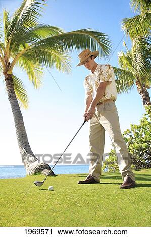 Stock Image - Hawaii, Oahu, Caucasion Male Standing And Posing With A Golf Club And Golf Ball In Hand At A Beautiful Tropical Beachfront Golf Course Turf. Fotosearch
