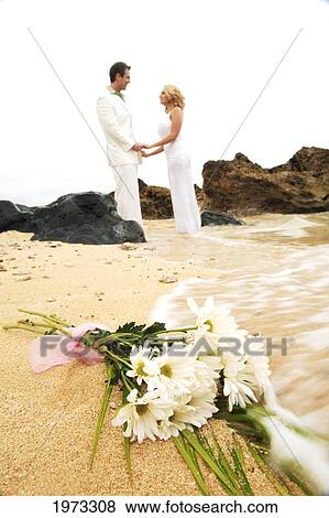 Stock Photo - Hawaii, Oahu, Caucasion Wedding Couple Holding And Looking Into Each Other With Bouquet Of White Daisies In Foreground Being Washed Up By The Ocean At The Beach. Fotosearch