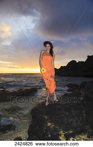 Stock Photograph - Hawaii, Oahu, Eternity Beach, Beautiful young woman in an orange dress holds a yellow hibiscus flower.. Fotosearch