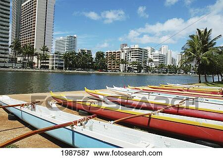 Stock Photo - Hawaii, Oahu, Honolulu, Lineup Of Colorful Outrigger Canoes Next To Ala Wai Canal, Waikiki Buildings Across The Water. Fotosearch