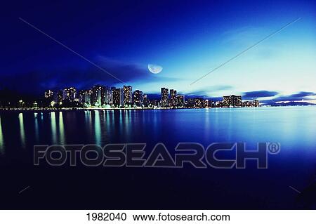 Stock Image - Hawaii, Oahu, Honolulu, Waikiki skyline at night, moon overhead, view from ocean. Fotosearch