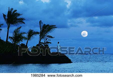 Picture - Hawaii, Oahu, Kahala Coastline, Woman Standing On Rocks Watching Moon Rise Over Blue Ocean, View From Behind.. Fotosearch