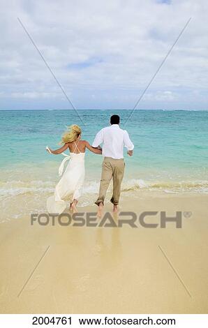 Stock Image - Hawaii, Oahu, Kailua, Lanikai Beach, Attractive Newlywed Couple Running Toward Ocean, View From Behind.. Fotosearch