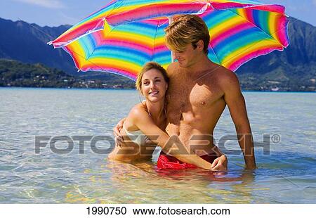 Stock Image - Hawaii, Oahu, Kaneohe, Couple Under A Brightly Colored Umbrella In Crystal Clear Water At The Sandbar Or Dissapearing Island. Fotosearch