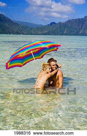 Stock Photograph - Hawaii, Oahu, Kaneohe, Couple Under A Brightly Colored Umbrella In Crystal Clear Water At The Sandbar Or Dissapearing Island. Fotosearch
