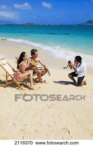 Stock Image - Hawaii, Oahu, Lanikai, A beachboy takes a picture of a young couple on the beach.. Fotosearch