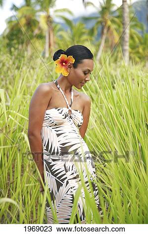 Stock Image - Hawaii, Oahu, Local Female In Aloha Hawaiian Attire Dress Standing In A Coconut Tree Field With Tall Green Grass. Fotosearch