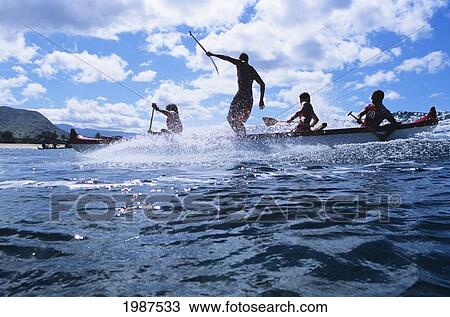 Stock Image - Hawaii, Oahu, Makaha, Man Standing Up In Outrigger Canoe While Surfing Wave, View From Side. No Model Release. Fotosearch