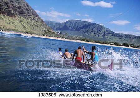 Stock Photograph - Hawaii, Oahu, Makaha, Paddlers In Outrigger Canoe Surfing Wave, View From Behind, Mountains In Background. No Model Release. Fotosearch