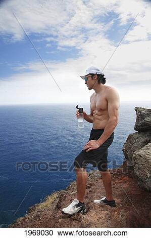 Stock Image - Hawaii, Oahu, Makapuu, Athletic Male Holding Water Bottle Taking A Break At Makapuu Cliffs. Fotosearch