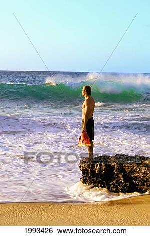 Stock Photograph - Hawaii, Oahu, Sandy Beach, Bodysurfer Stands On Rocks Looking At Ocean, Holding Fins.. Fotosearch