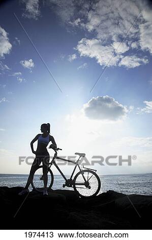 Stock Image - Hawaii, Oahu, Silhouette Of A Woman With A Bike On A Rocky Coastline.. Fotosearch