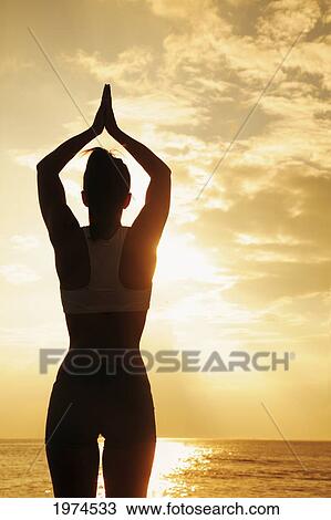 Stock Image - Hawaii, Oahu, Silhouette Of Beautiful Girl Standing On The Rocky Coastline As The Sun Sets With Boat In Background.. Fotosearch