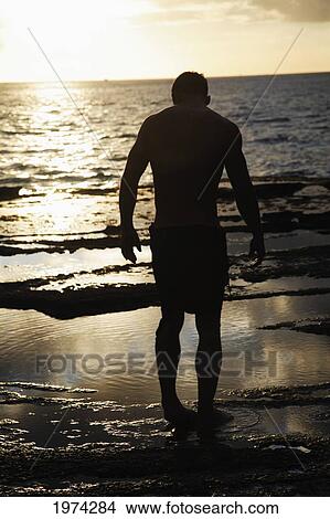 Picture - Hawaii, Oahu, Silhouette Of Fit Man Standing On The Rocky Coastline As The Sun Sets.. Fotosearch