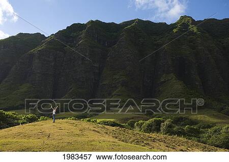 Stock Photo - Hawaii, Oahu, Teenage Girl On A Hill In The Distance Raising Arms To The Sky, Dramatic Mountain Landscape.. Fotosearch