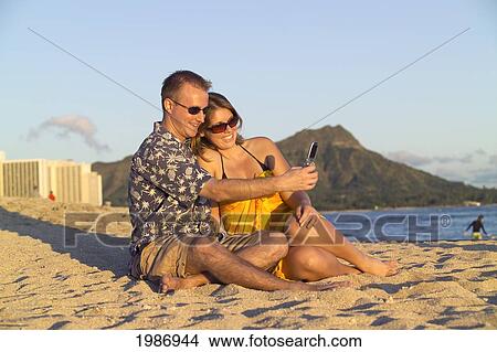 Picture - Hawaii, Oahu, Waikiki Beach, Couple Sitting On The Beach With Diamond Head In The Background, Take Photo Of Themselves With A Camera Phone.. Fotosearch