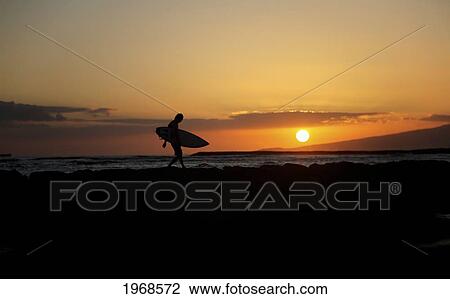 Stock Image - Hawaii, Oahu, Waikiki, Silhouette Of A Surfer Walking On A Rock Wall At Sunset. Fotosearch