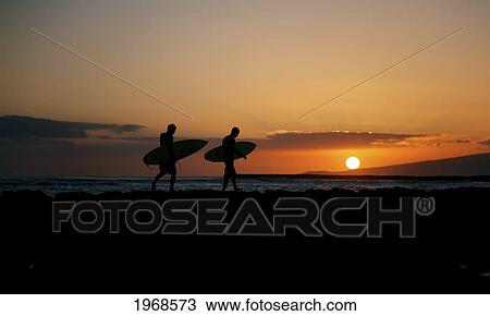 Stock Image - Hawaii, Oahu, Waikiki, Two Surfers Walking Along The Shoreline As The Beautiful Sunsets. Fotosearch