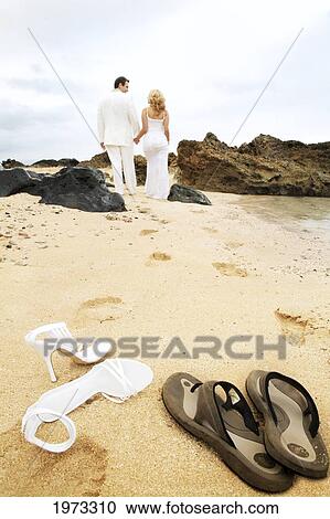 Stock Image - Hawaii, Oahu, Wedding Couple Walking Along Beach Leaving Behind Footprints In The Sand And Their Footwear In Foreground, View From Behind.. Fotosearch