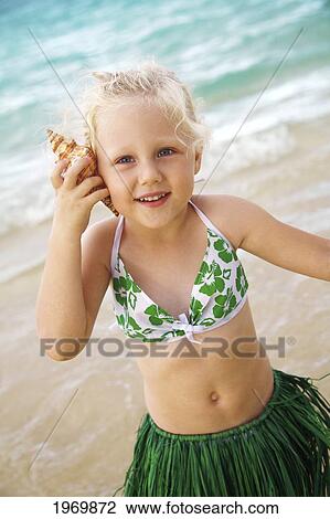 Stock Image - Hawaii, Oahu, Young Girl In A Hula Skirt Listening To A Shell.. Fotosearch