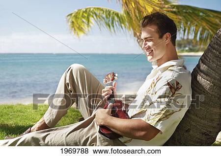 Stock Photo - Hawaii, Oahu, Young Male With Vintage Aloha Shirt Relaxing On Palm Tree, Holding A Ukulele.. Fotosearch