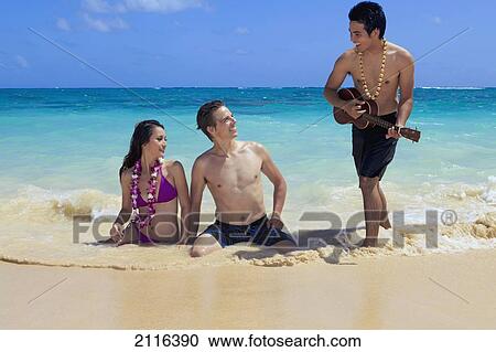 Stock Image - Hawaii, polynesian man plays ukulele for a young couple in the ocean.. Fotosearch
